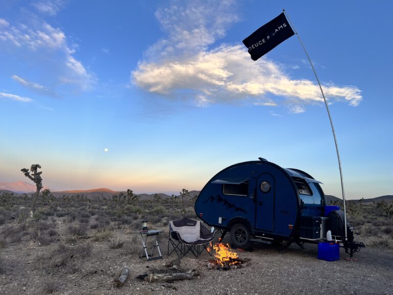 Off-grid campsite at sunset with a teardrop camper, campfire, and a flag reading “Deuce Roams” waving above the desert landscape.