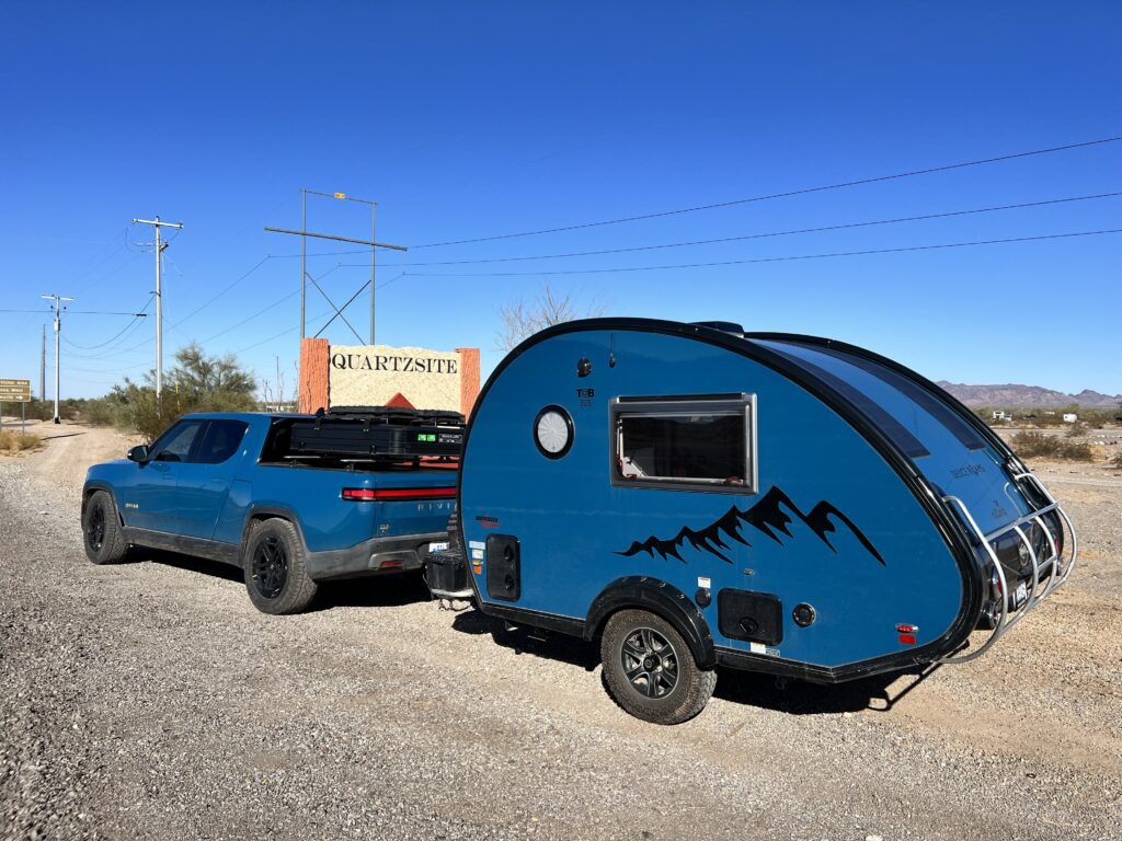 Blue Rivian R1T with matching teardrop trailer parked at the Quartzsite welcome sign in the Arizona desert while on an adventure with purpose..