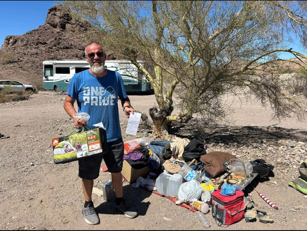 Jeremy holding a new tent and receipt next to belongings of an unhoused individual in the desert near an RV