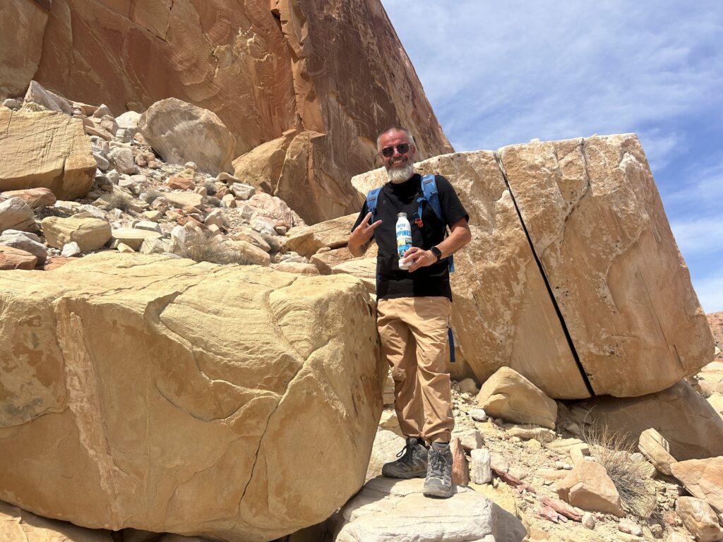 Jeremy standing on desert rocks with a backpack and water bottle, surrounded by towering sandstone cliffs under a partly cloudy sky.