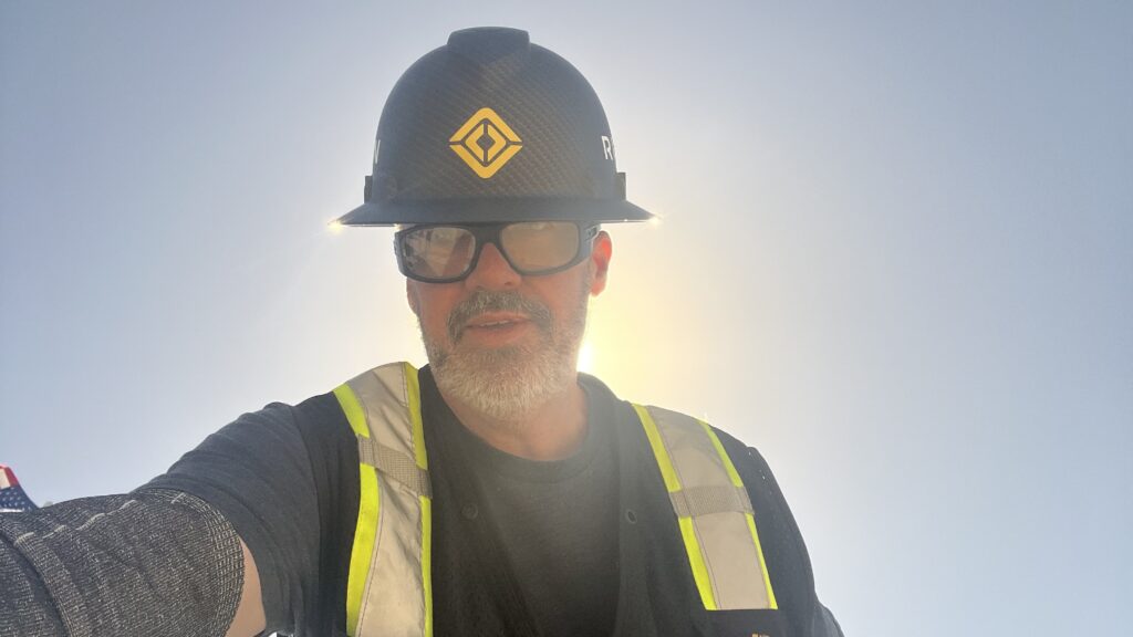 Jeremy wearing a Rivian hard hat and reflective vest, standing in front of the sun with a soft glow surrounding him.