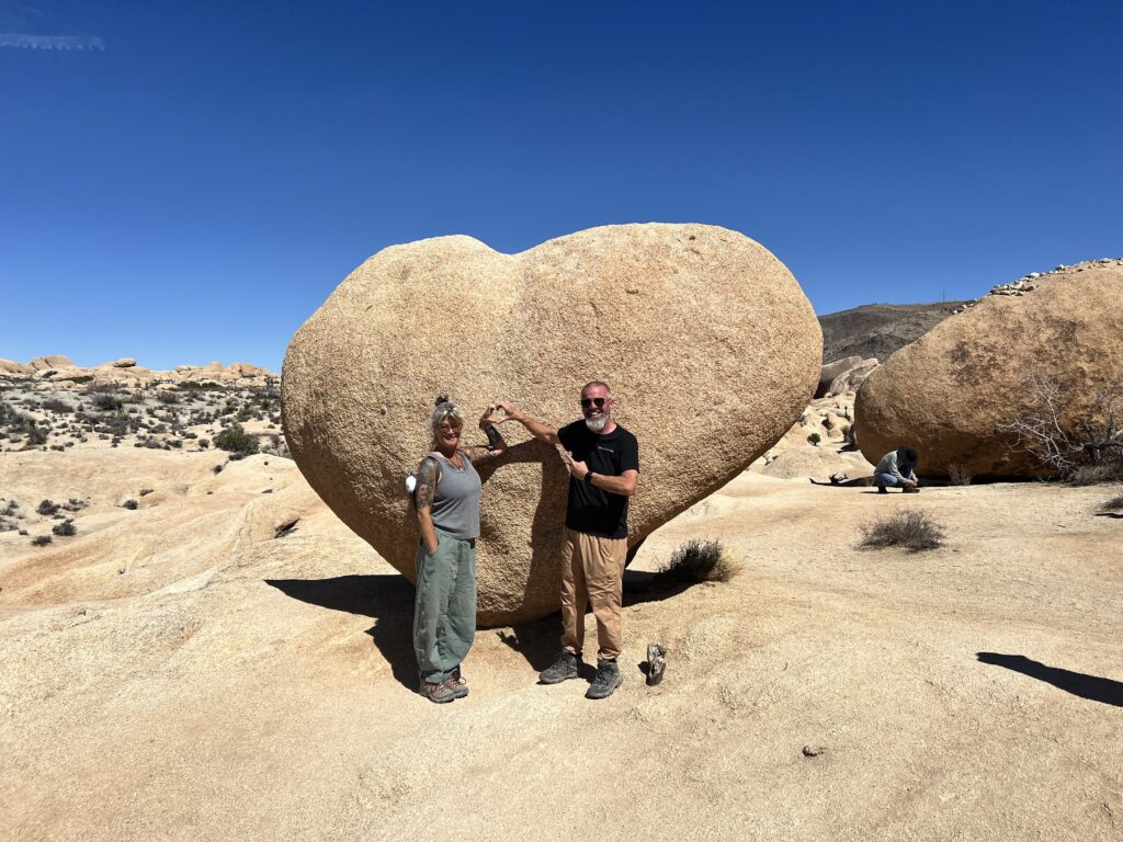 Two nomads forming a heart with their hands in front of Heart Rock in Joshua Tree National Park as they adventure with purpose.