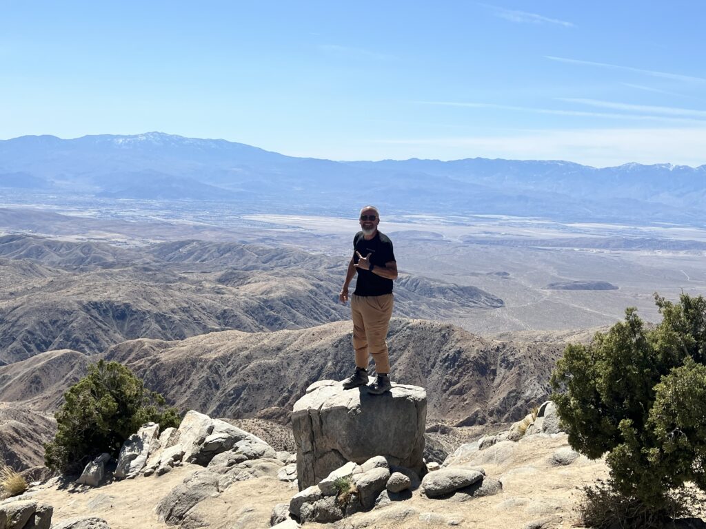 Jeremy standing on a large rock at the edge of a mountain viewpoint, overlooking a vast desert valley under a clear blue sky.