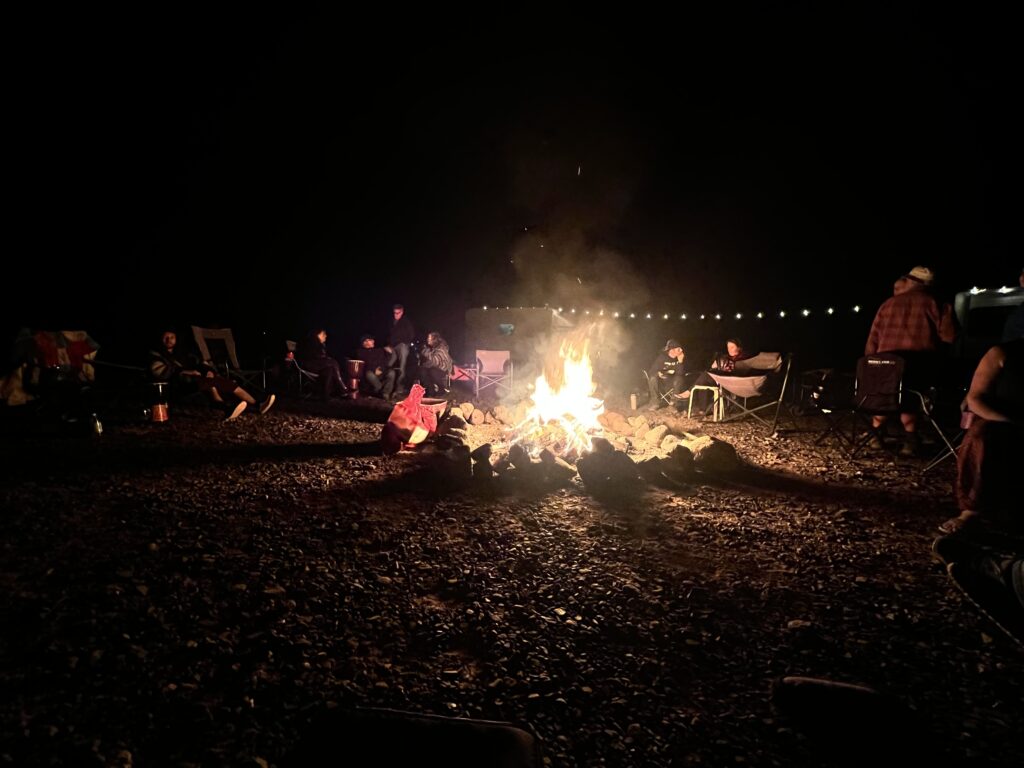 A group of people gathered around a glowing campfire at night, seated in a circle with string lights in the background.