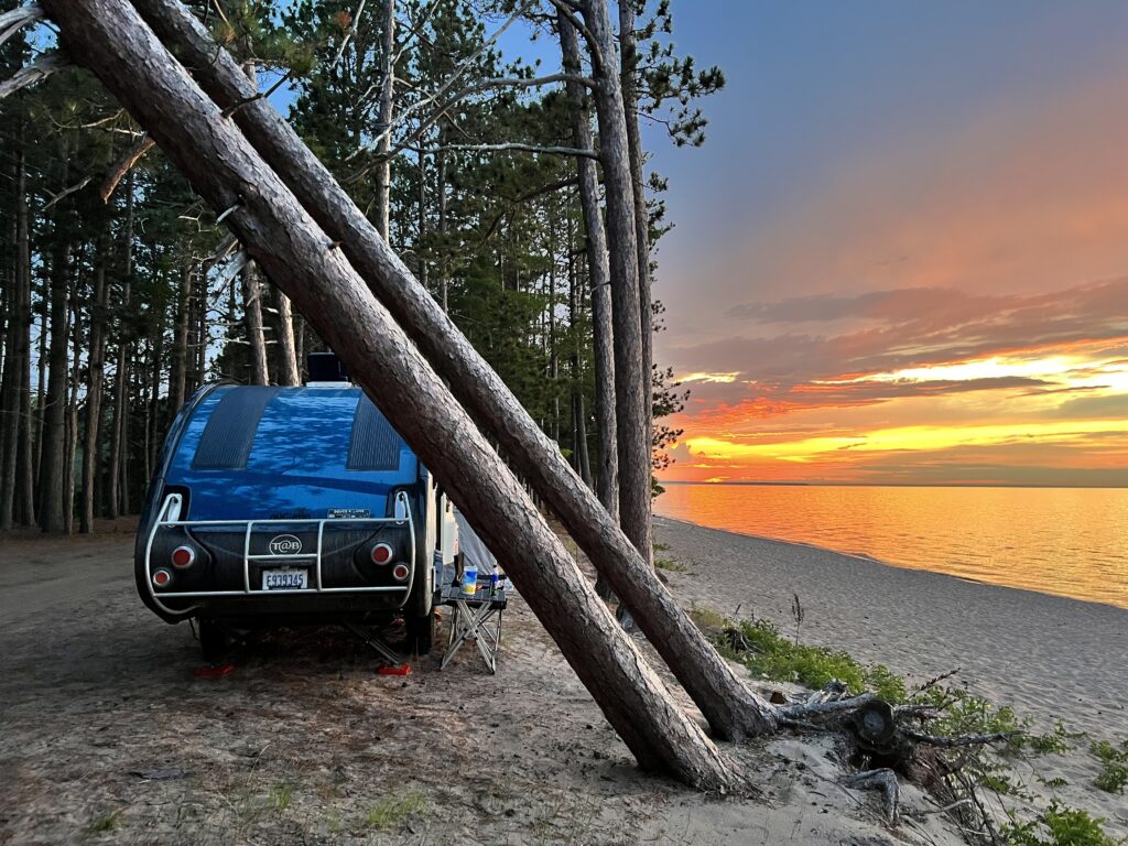 NuCamp Tab 320s camper parked beside Lake Superior at sunset with vibrant orange skies and leaning pine trees