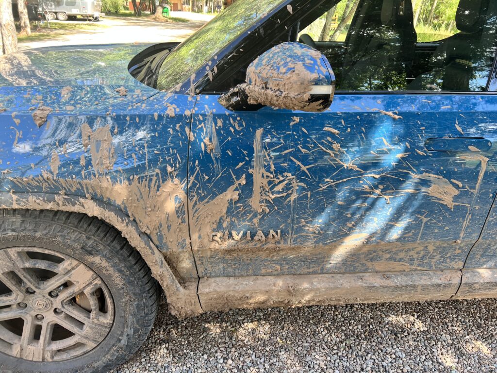 Close-up of a mud-covered Rivian R1T, parked on gravel after an off-road adventure.