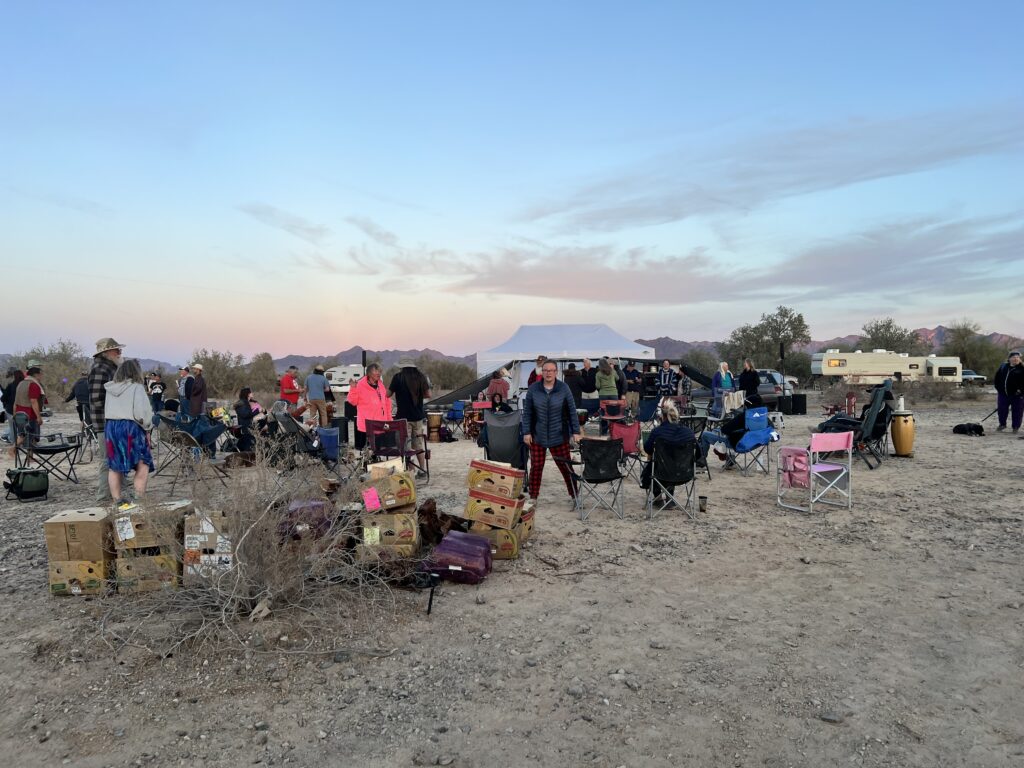 Nomads gathered in the desert around a community tent at sunset, surrounded by chairs, supplies, and RV's.