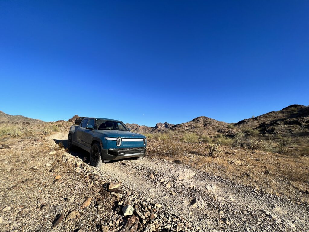 Electric truck parked on a rugged desert trail under a clear blue sky, surrounded by rocky terrain and distant mountains.