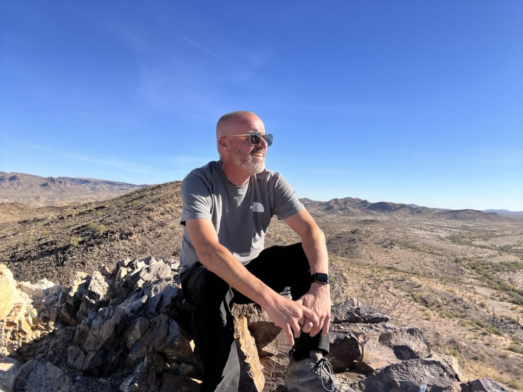 Jeremy sitting on a rocky summit overlooking the desert, reflecting on his adventure with purpose under a clear blue sky.