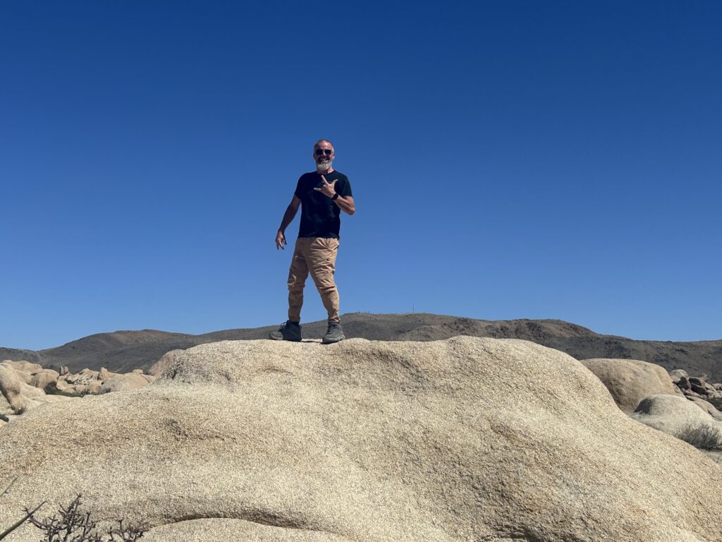 Jeremy standing on large desert boulder under blue sky, symbolizing freedom and peace from nomadic travel.
